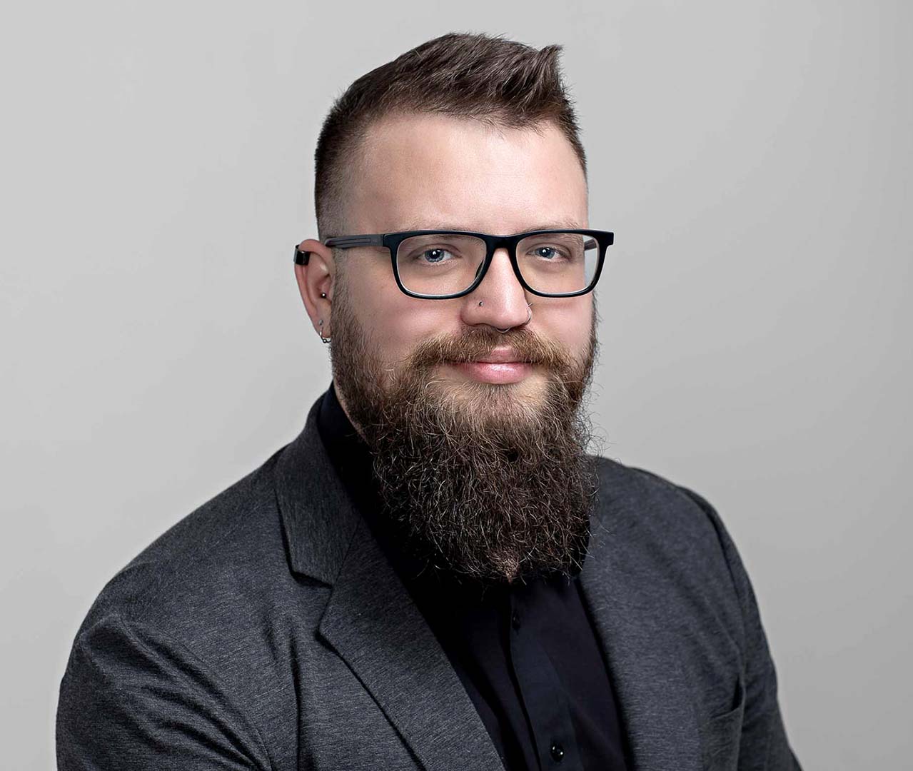 young man with beard poses for an executive portrait wearing black glasses, grey suit and black shirt