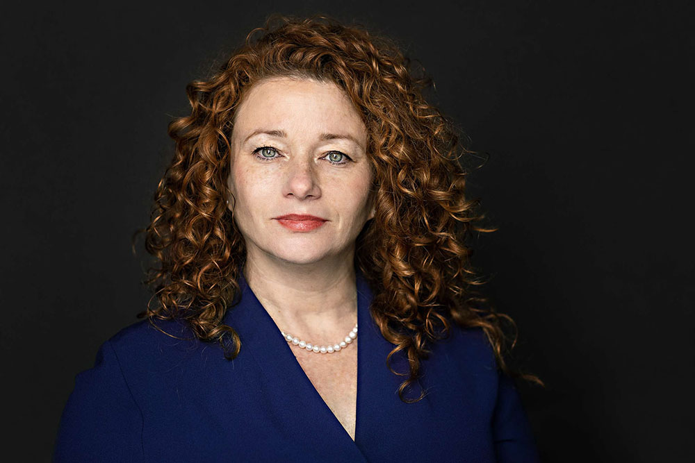 woman with red hair and blue dress poses for a corporate executive headshot on dark background