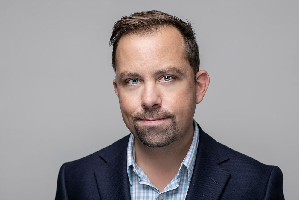 man in blue check shirt poses for a corporate executive headshot against grey background