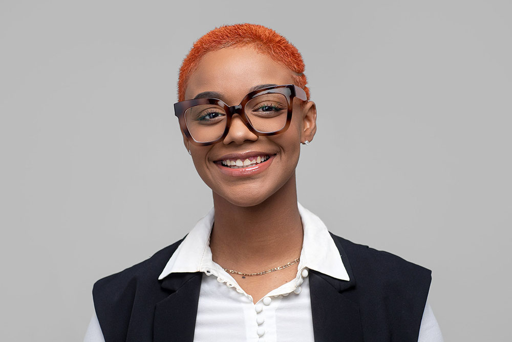 woman with orange hair and large glasses poses for corporate headshot against grey