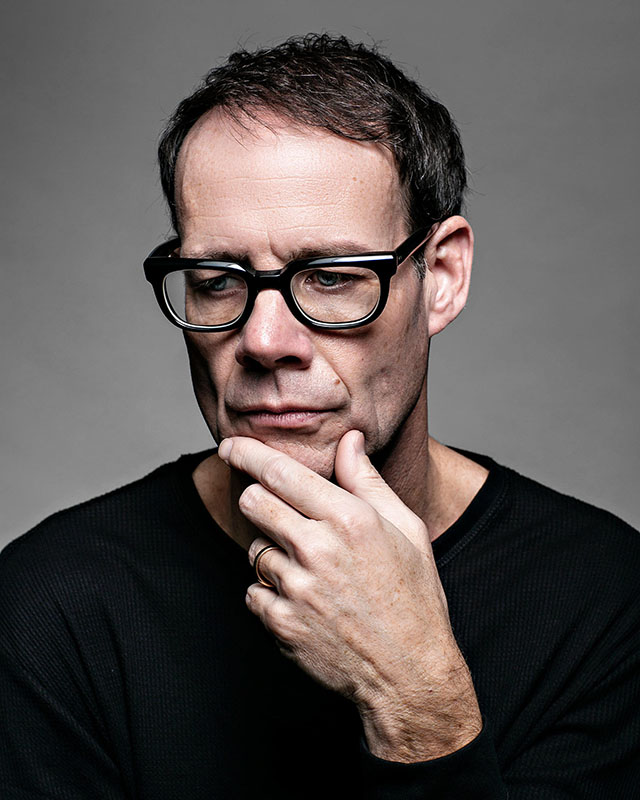actor wearing black shirt and glasses posing for a headshot portrait on grey background